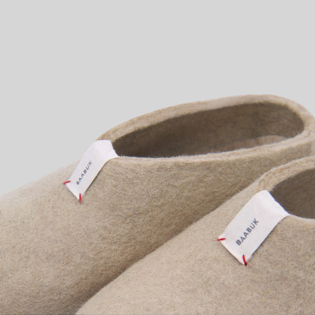 Close-up of a pair of beige wool felt slip-on slippers with white fabric labels reading BAABUK and small red stitched accents, photographed on a light gray background.