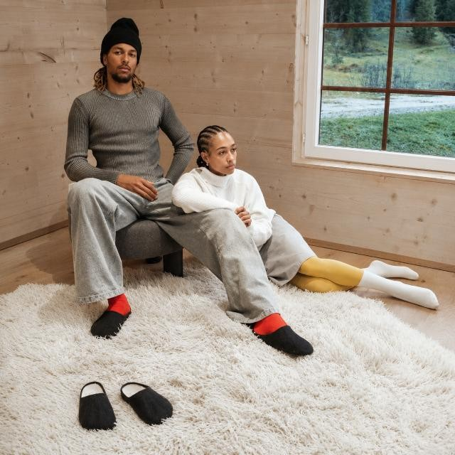 Baabuk Alpina black wool slip-on sandals worn by a man and woman in a cozy wooden room setting