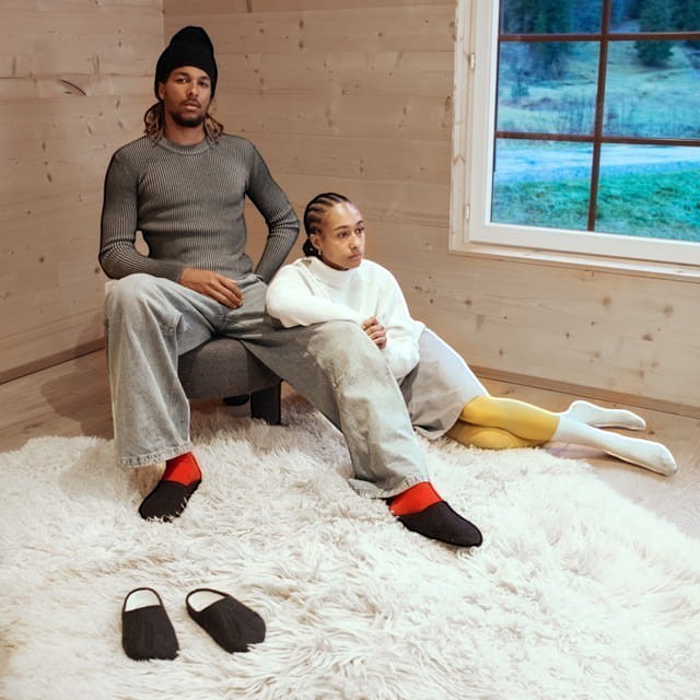 Baabuk Alpina black wool slip-on sandals worn by models in a cozy wooden cabin setting.