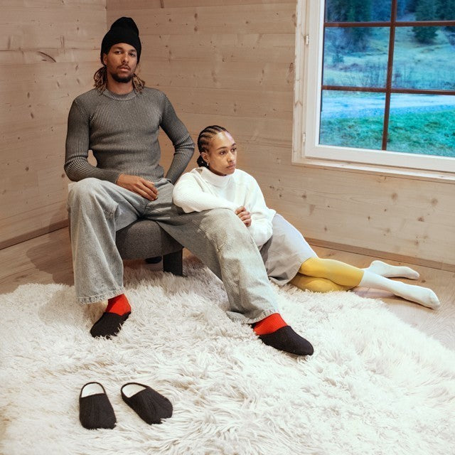 Baabuk Alpina black wool slip-on sandals worn by a couple in a cozy wooden cabin setting.