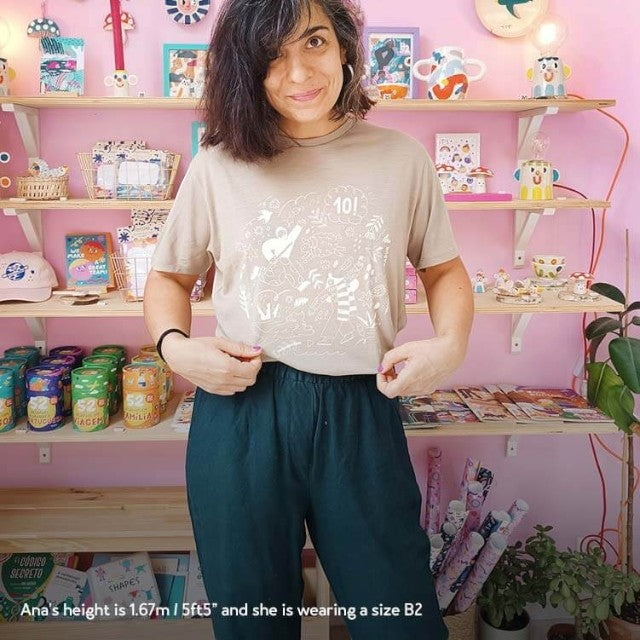 A woman wearing a khaki Baabuk 10th anniversary merino wool t-shirt with a white illustration in a colorful studio.
