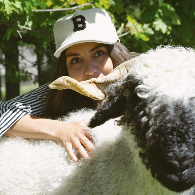 Baabuk B Cap in off-white wool with black embroidered B, worn by a woman next to a Valais Blacknose sheep.