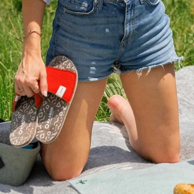 Baabuk Baabulette wool slide sandals in Swiss Red with patterned footbeds held by a woman outdoors on a picnic blanket