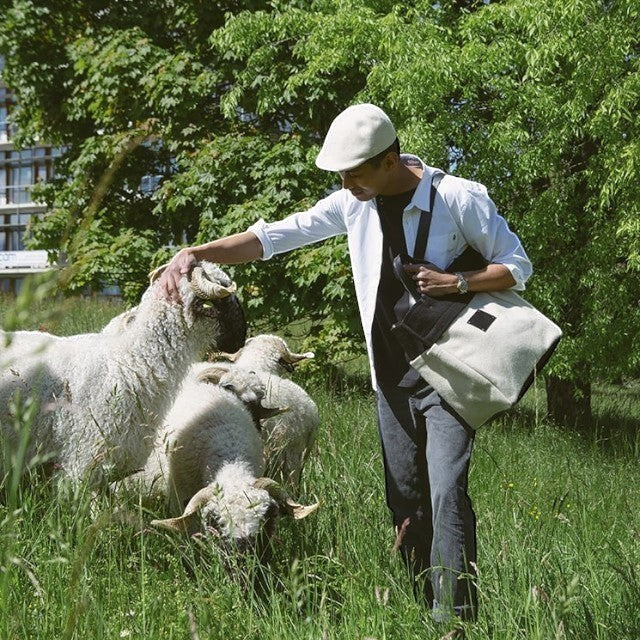 Man wearing a Baabuk BBB Bag in a field with Valais Blacknose sheep, featuring a two-tone wool tote with leather.