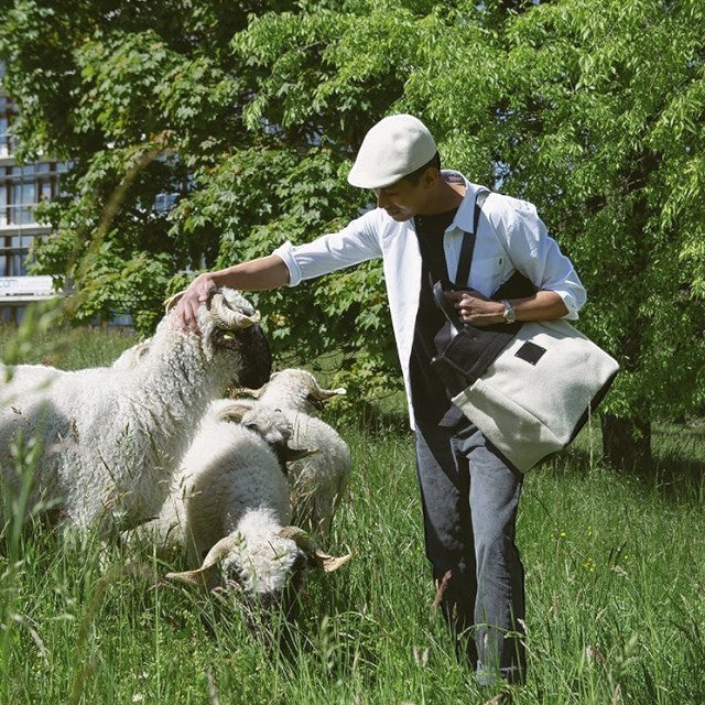 Man wearing a Baabuk BBB Bag in a field with Valais Blacknose sheep, featuring a two-tone wool tote with leather.