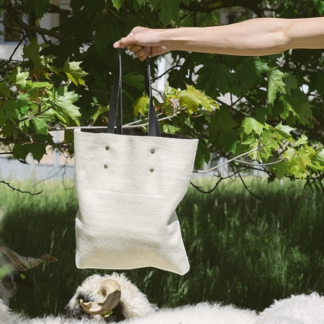 Baabuk Blacknose reversible tote bag made from cream Swiss wool and leather, held outdoors near a sheep.