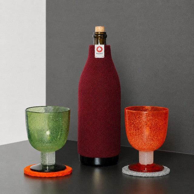 Baabuk bordeaux wool bottle cover insulating a wine bottle next to green and orange textured glasses on a table