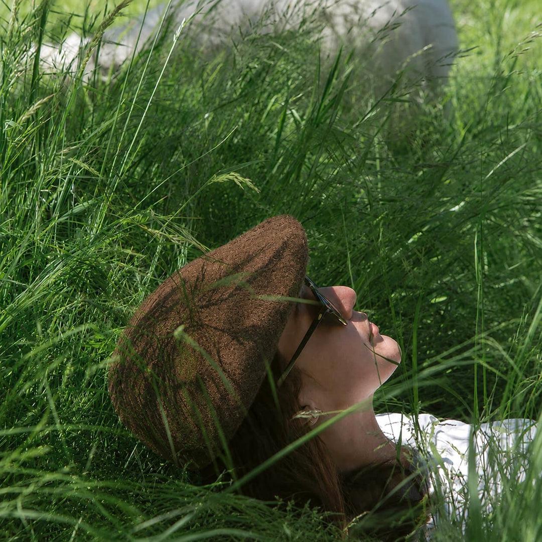 Baabuk brown wool beret worn by a person lying in tall green grass, inspired by traditional Portuguese shepherd hats.