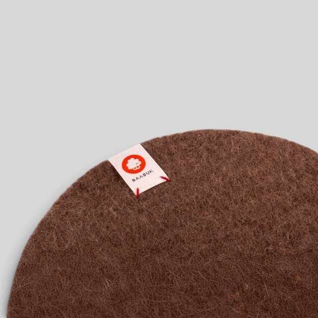 Brown Baabuk felted wool trivet hot plate with a small branded fabric tag, perfect for heat-resistant table protection.