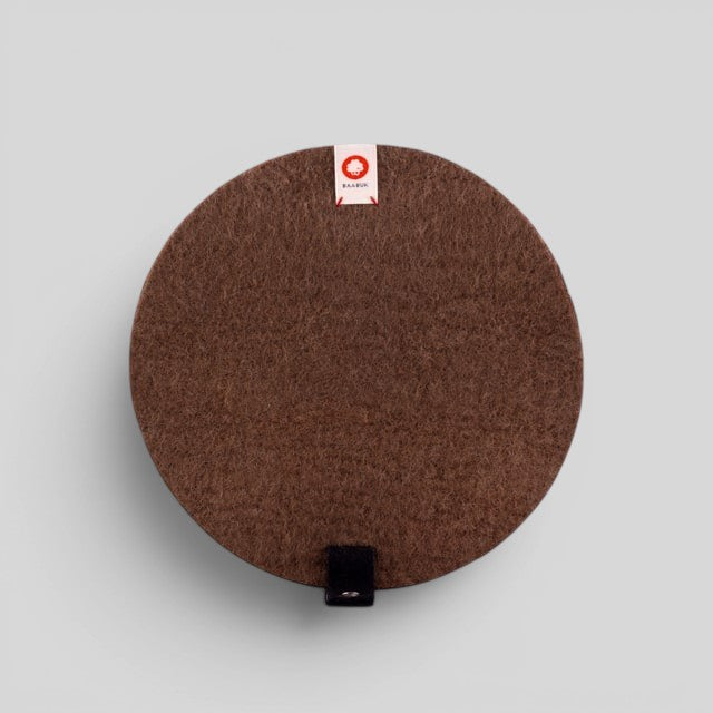 Baabuk round brown felted wool trivet hot plate with a small leather hanging strap and branded fabric tag.