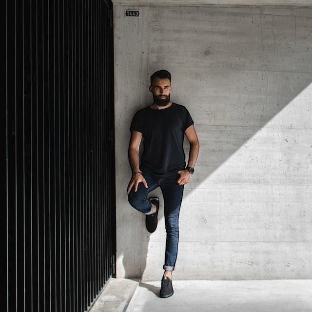 Baabuk Classic Wooler black sneakers worn by a bearded man in a dark t-shirt and jeans leaning against a concrete wall