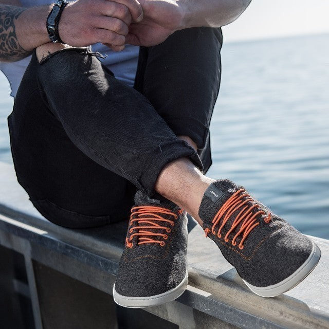 Baabuk Classic Wooler sneakers in grey wool with bright orange laces worn by a man sitting near the water.