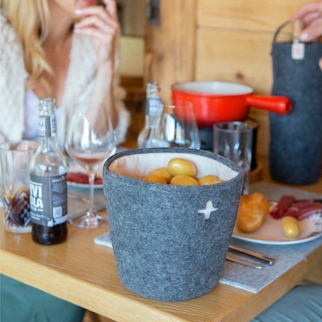 Baabuk dark grey felted wool basket with Swiss cross detail, filled with potatoes on a wooden dining table.