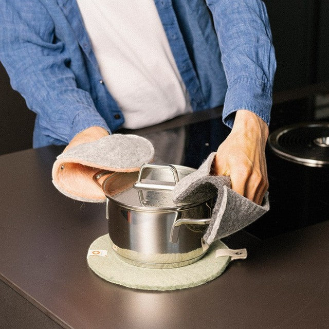 Person using dark grey Baabuk felted wool potholders to lift a stainless steel pot off a kitchen counter.