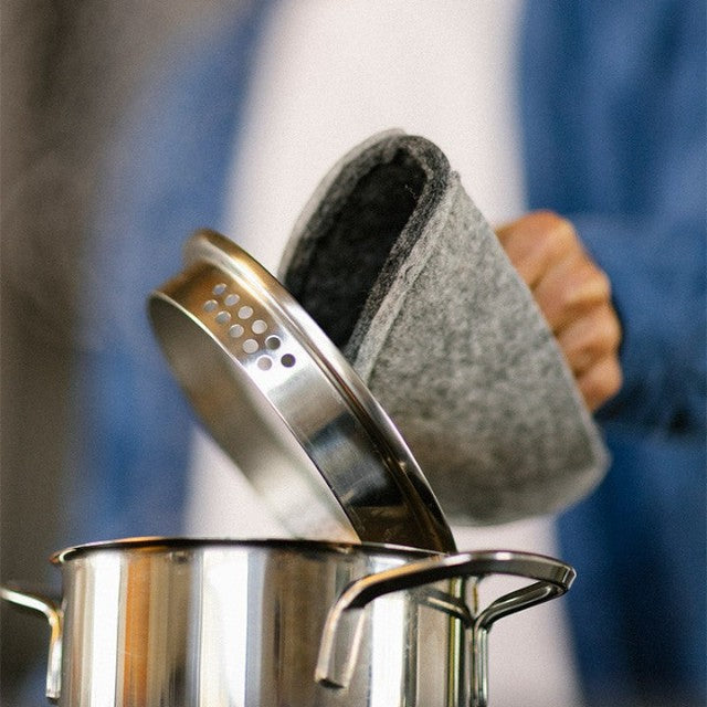 Baabuk dark grey felted wool potholder with leather strap, used to lift a hot stainless steel pot lid in the kitchen.