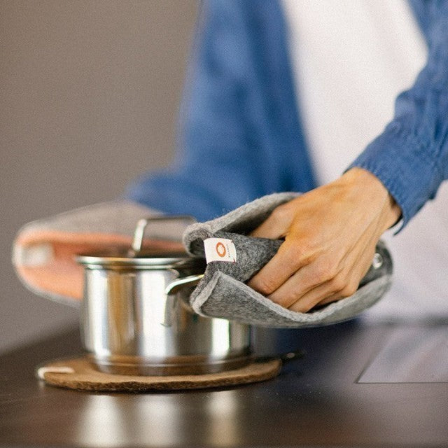 Baabuk dark grey felted wool trivet under a stainless steel pot on a table, held by a hand in a matching mitt.