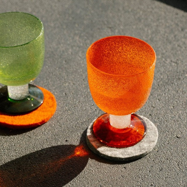 Baabuk khaki felted wool coasters under vibrant orange and green textured glass goblets on a grey surface.