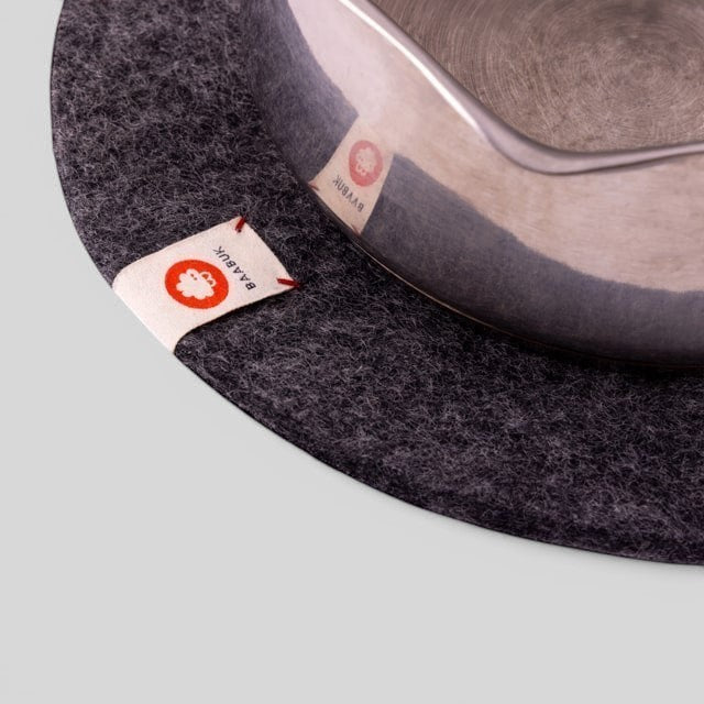 Baabuk dark grey felted wool trivet hot plate with a leather strap detail under a metal pot on a neutral background.