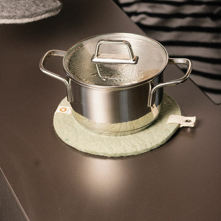 Baabuk round felted wool trivet in light green under a stainless steel pot on a dark kitchen countertop