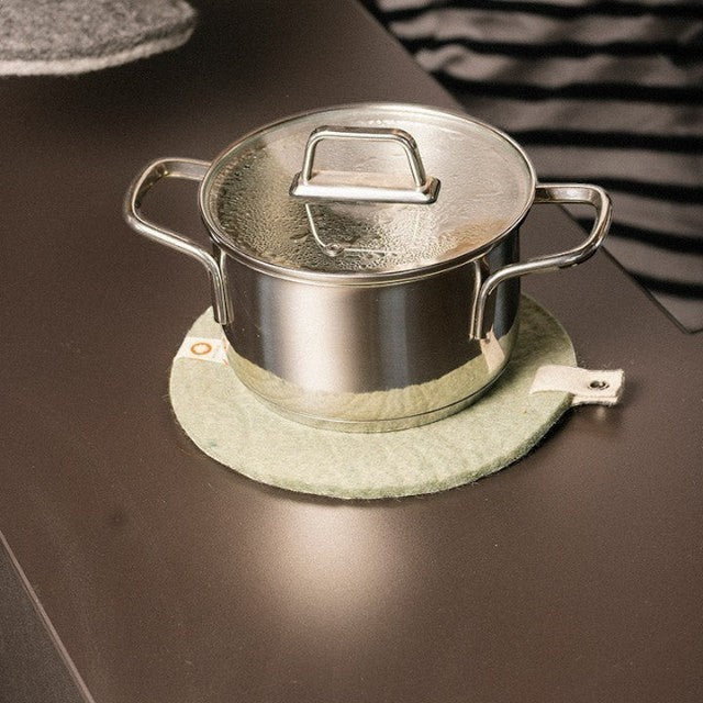 Baabuk round felted wool trivet in light grey protecting a dark countertop under a stainless steel cooking pot.