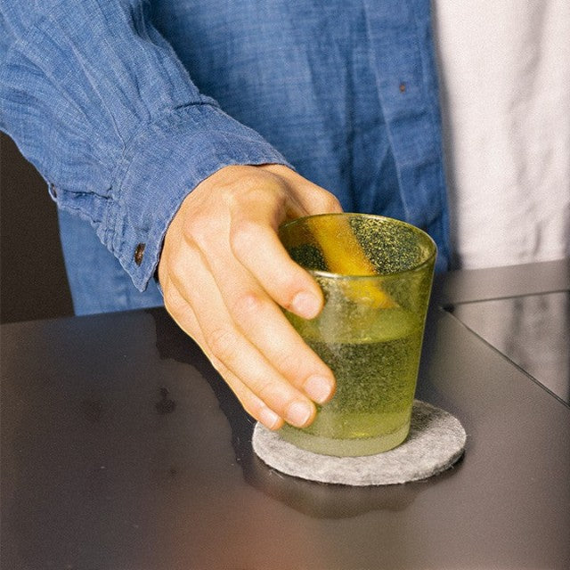 Baabuk khaki felted wool coaster under a glass held by a person wearing a blue shirt on a dark table.