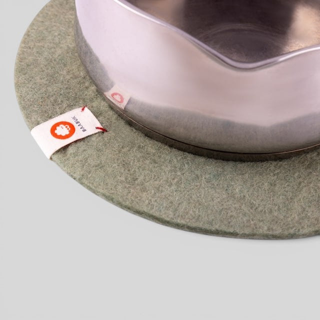 Baabuk khaki felted wool trivet hot plate with a metal pot on top, featuring a branded fabric tag.