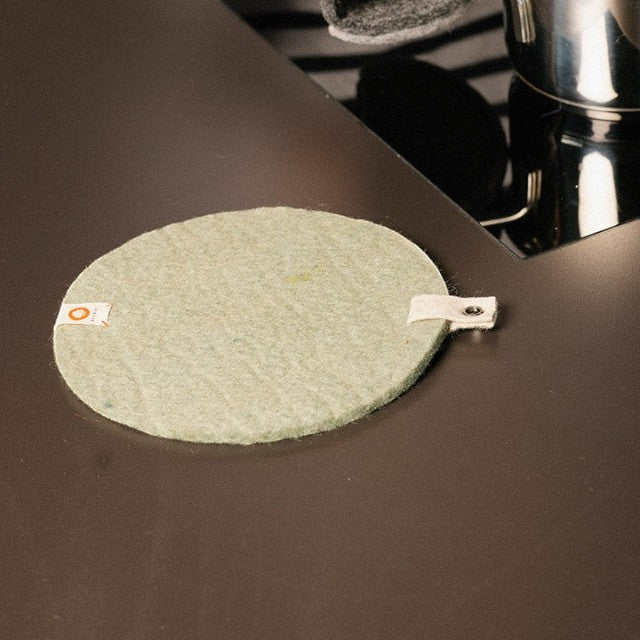 Baabuk khaki felted wool trivet hot plate with a small side strap on a dark kitchen countertop.