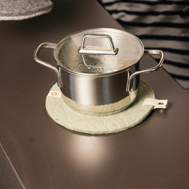 Baabuk khaki felted wool trivet hot plate with a stainless steel pot on a dark kitchen countertop.
