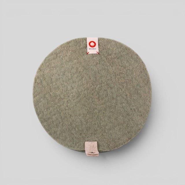Baabuk round khaki felted wool trivet hot plate with a small leather strap and branded label on a grey background.
