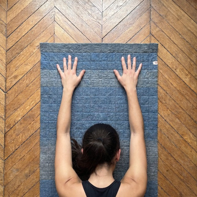 Baabuk light blue felted wool yoga mat with cotton stitching, shown from above with a person in a yoga pose.