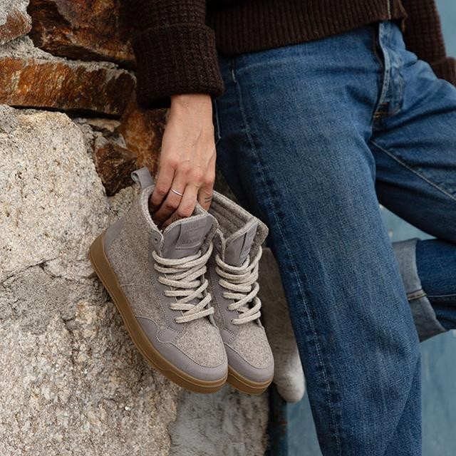 Baabuk Peaks Wooler sneakers in warm grey wool held by a person wearing jeans against a stone wall.