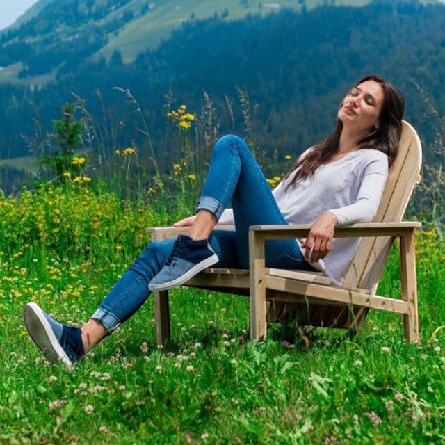 Woman relaxing in a wooden chair wearing Baabuk Sky Wooler blue wool sneakers in a scenic mountain meadow.