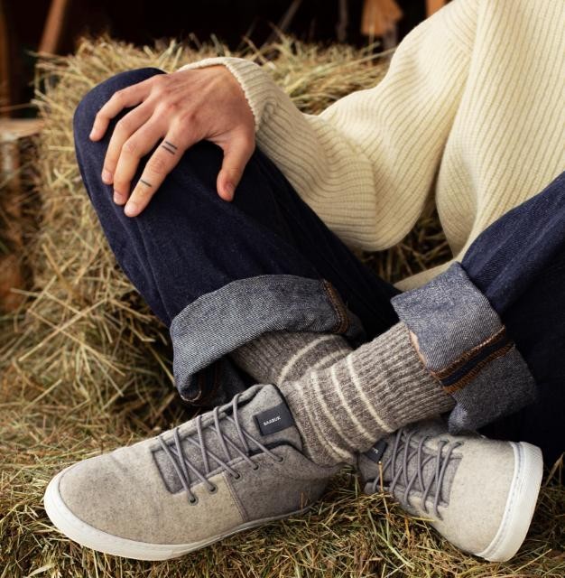 Baabuk Sky Wooler sneakers in middle grey wool with rubber soles, worn by a person sitting on hay in dark jeans.