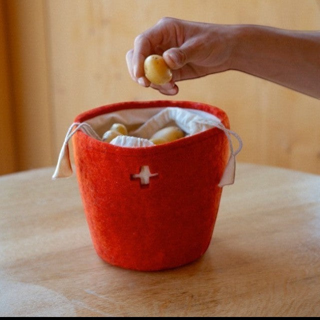 Baabuk Swiss red felted wool basket with a cross cutout and drawstring, holding potatoes on a wooden table.