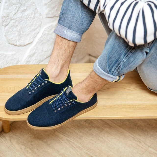 Baabuk Urban Wooler sneakers in navy wool with yellow trim and elastic laces worn with jeans on a wooden bench