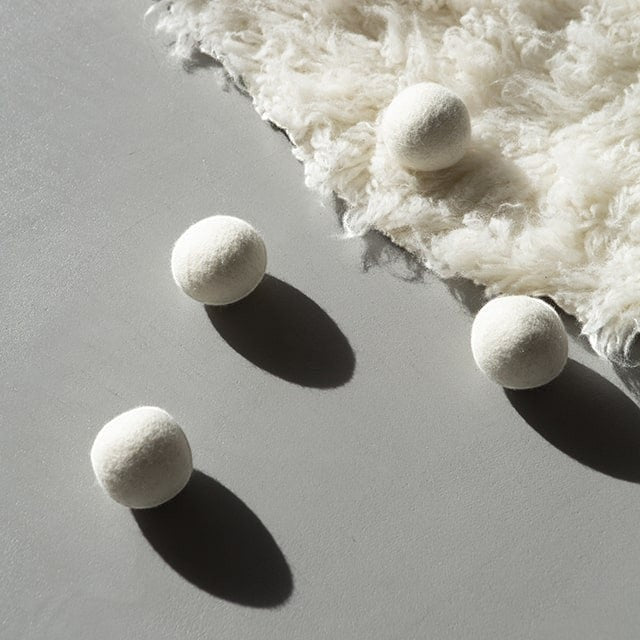 Four white Baabuk wool dryer balls on a grey surface next to a piece of raw sheep wool for sustainable laundry.