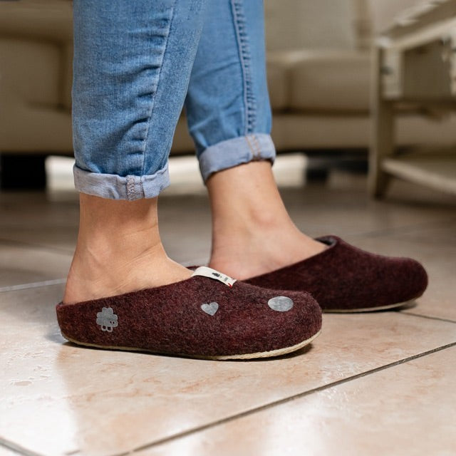 Baabuk wool felt slippers in dark burgundy with decorative patches worn with blue jeans on a tiled floor.