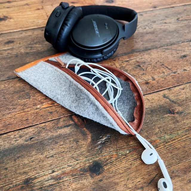 Baabuk grey wool zippered pouch with leather trim, holding white earbuds and black headphones on a wooden table.