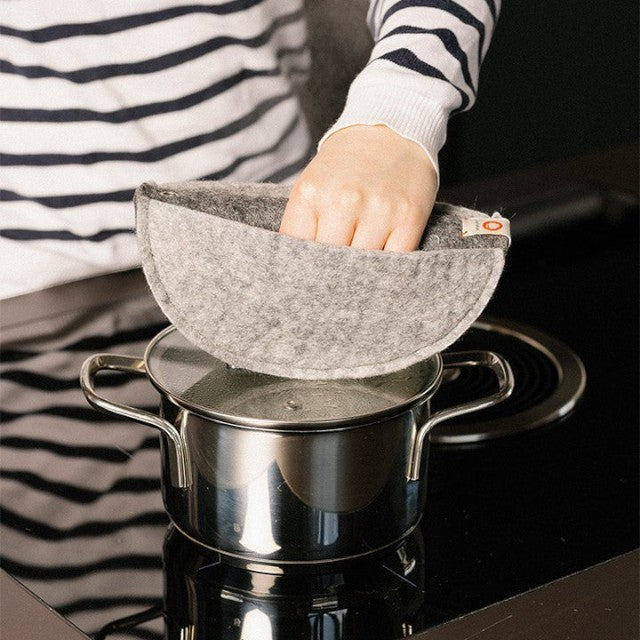 Baabuk grey felted wool potholder being used to lift a stainless steel pot lid on a black stovetop.