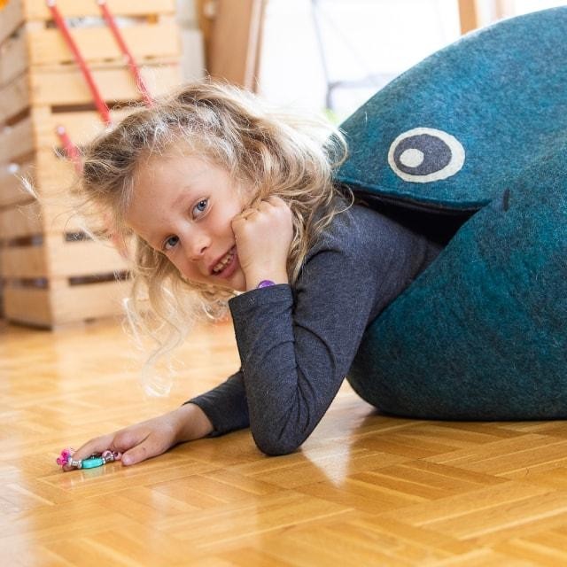 A young girl playing with a blue Baabuk handmade wool whale storage basket on a wooden floor.
