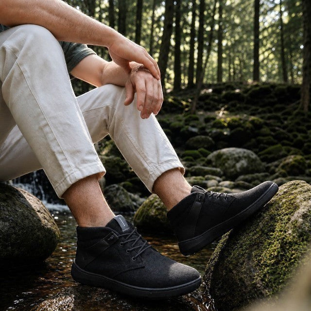 Baabuk Zero Wooler High black wool barefoot shoes worn by a person sitting on mossy rocks in a forest.